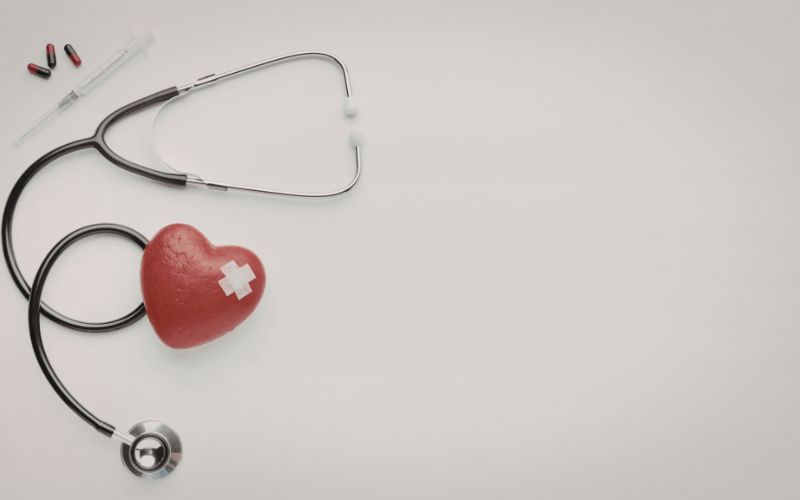 Stetoskop, czerwone serce z plastrem, strzykawka i tabletki na jasnym tle.